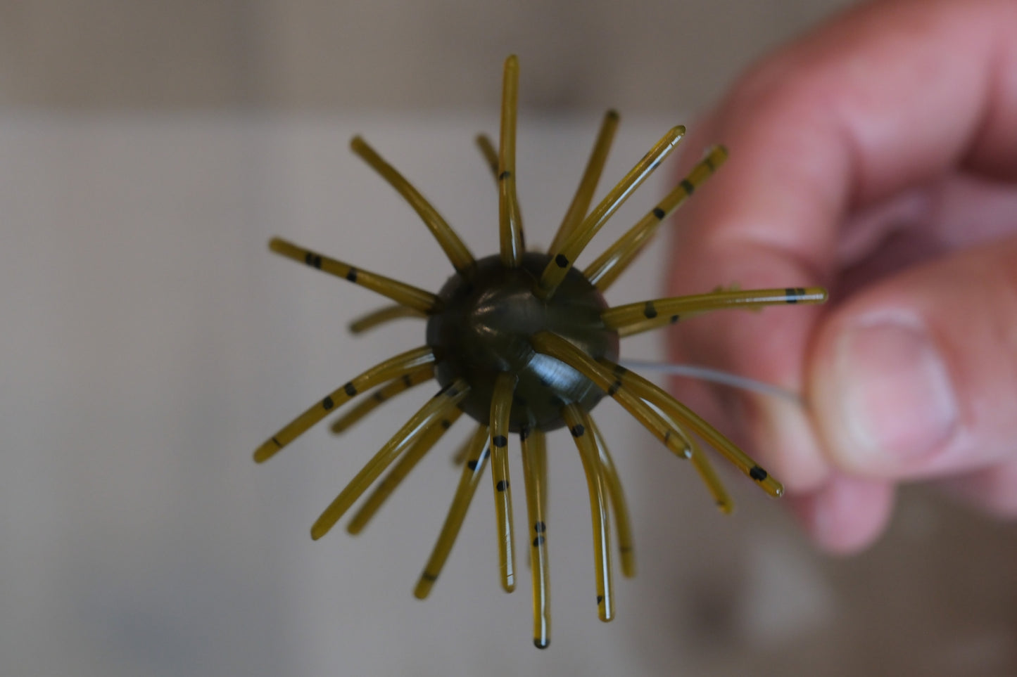 A close-up of a fishing lure being held by a hand. The lure has a dark olive-green center with multiple translucent yellow-brown legs tipped with small black rings, set against a blurred beige background.