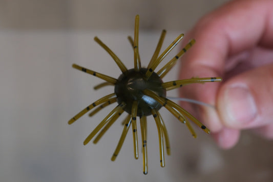 A close-up of a fishing lure being held by a hand. The lure has a dark olive-green center with multiple translucent yellow-brown legs tipped with small black rings, set against a blurred beige background.