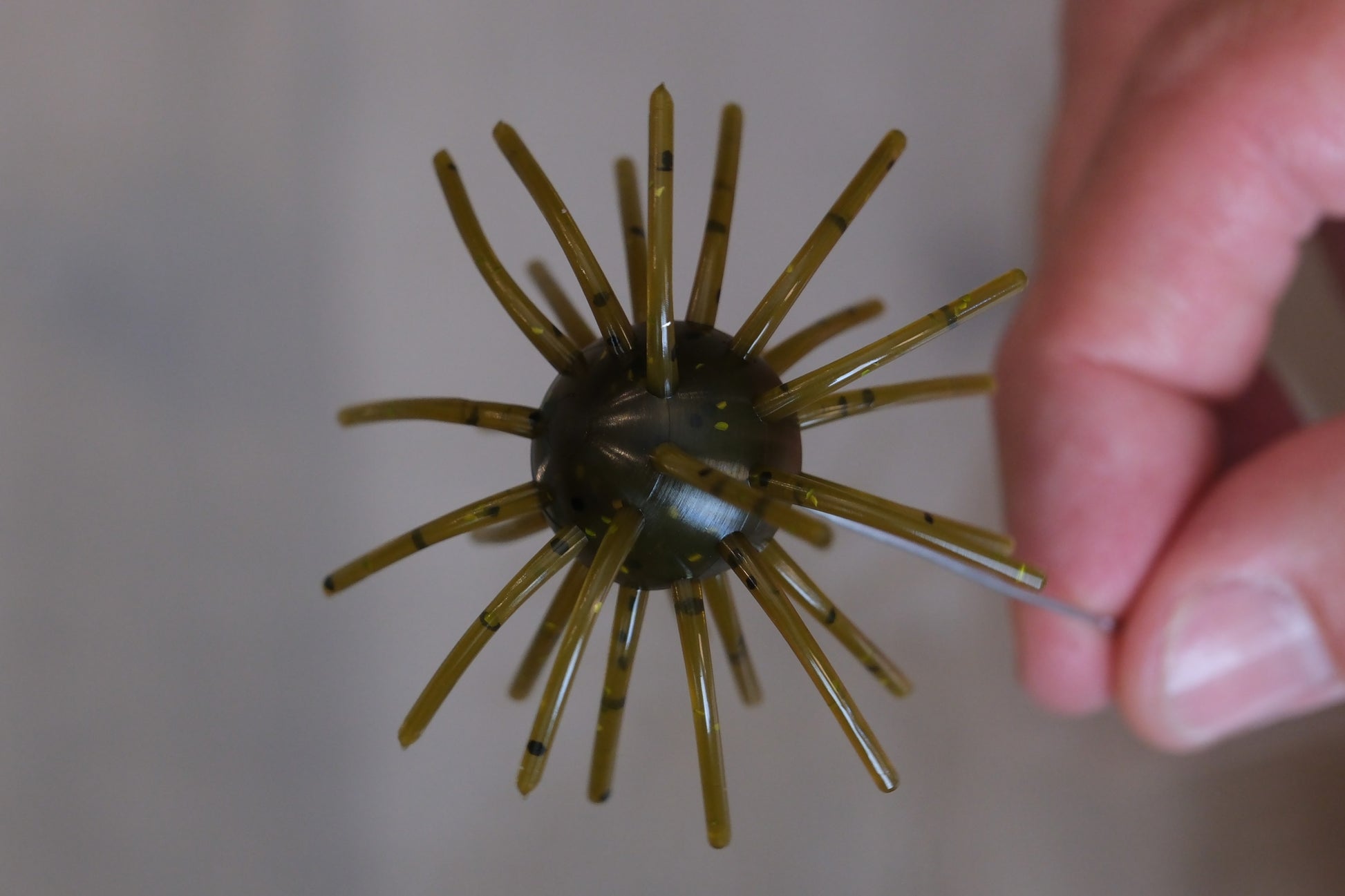 A close-up of a fishing lure with a dark center and numerous translucent olive-brown spines, held by a person's hand against a blurred gray background.
