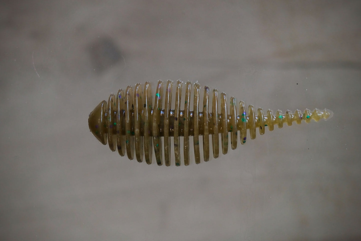 A small translucent fishing lure or bait with a ribbed, segmented body and iridescent flecks, shown against a plain light gray background.
