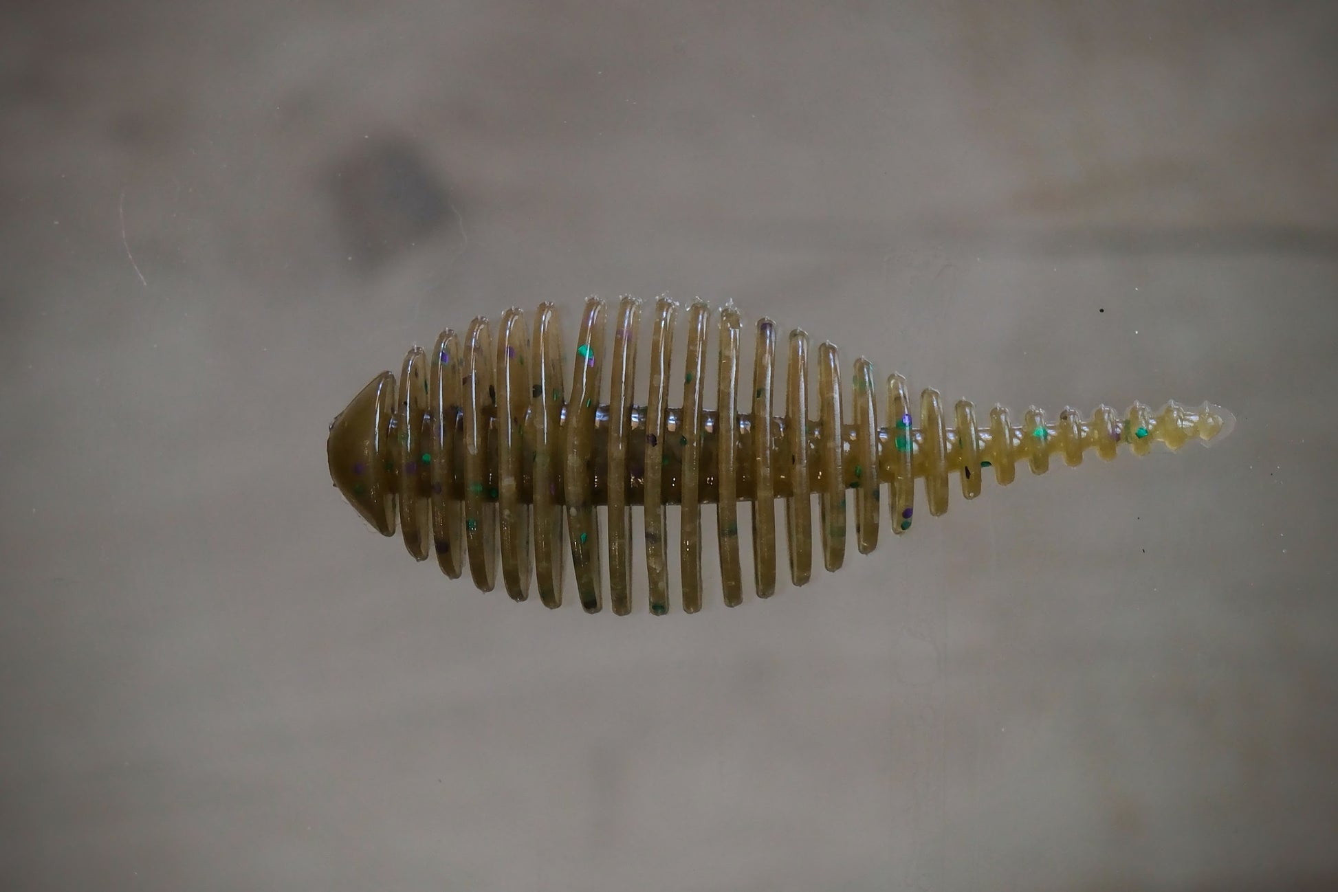 A small translucent fishing lure or bait with a ribbed, segmented body and iridescent flecks, shown against a plain light gray background.