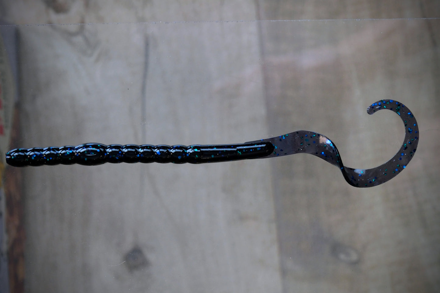 A curved hook-shaped fishing lure with a textured black handle and a glittery blue and black body, shown against a blurred wood background.