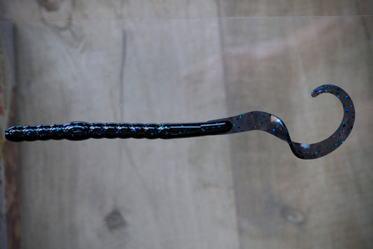 A curved hook-shaped fishing lure with a textured black handle and a glittery blue and black body, shown against a blurred wood background.