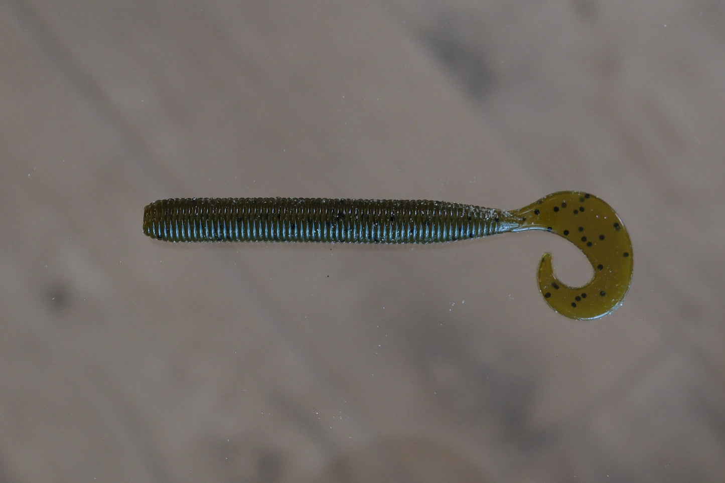 A soft plastic fishing lure shaped like a segmented worm with a curved tail, shown on a light gray surface.