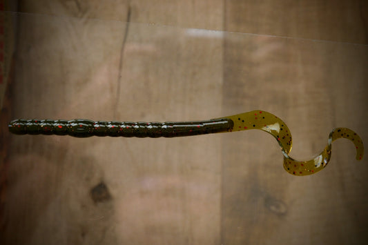 A soft plastic fishing lure with a ribbed dark olive-green body and a wavy tail in translucent yellow-green with black speckles, shown against a blurred brown wood-grain background.