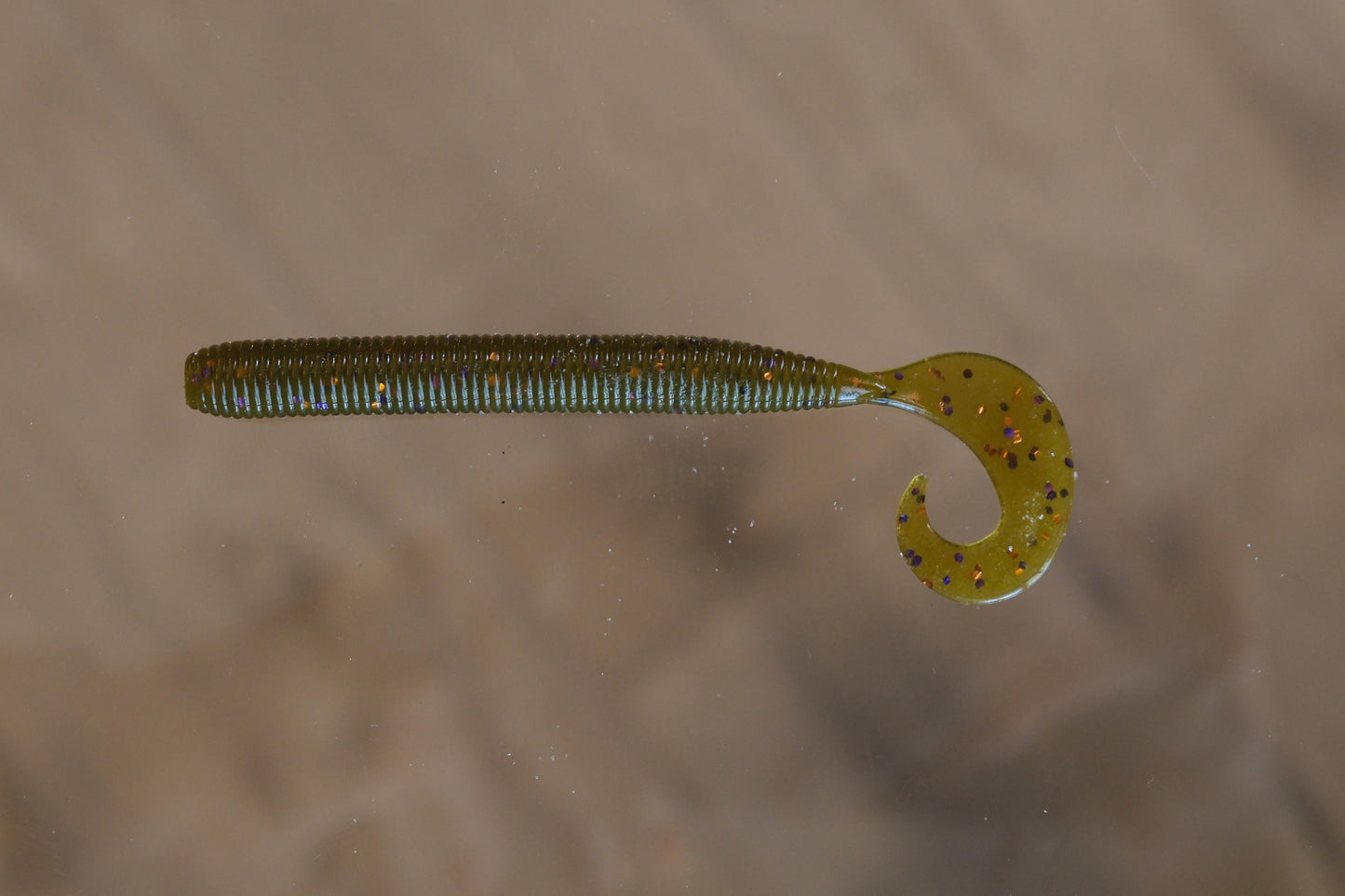 A soft plastic fishing lure shaped like a small segmented worm with a curved tail, shown against a blurred beige background.