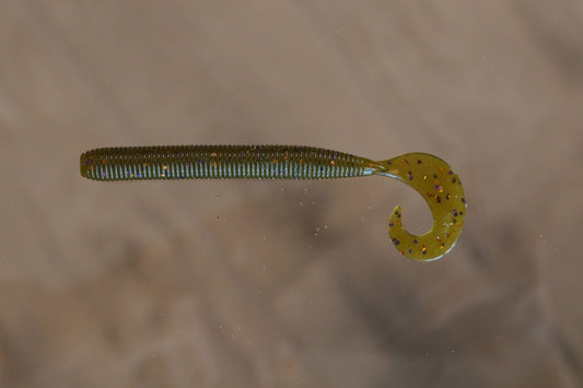 A soft plastic fishing lure shaped like a small segmented worm with a curved tail, shown against a blurred beige background.