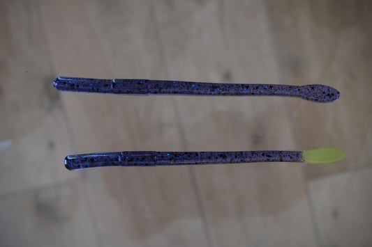 Two soft plastic fishing lures with a dark blue and black speckled pattern, displayed on a light wood floor. One lure has a dark tail, and the other has a bright chartreuse green tail.