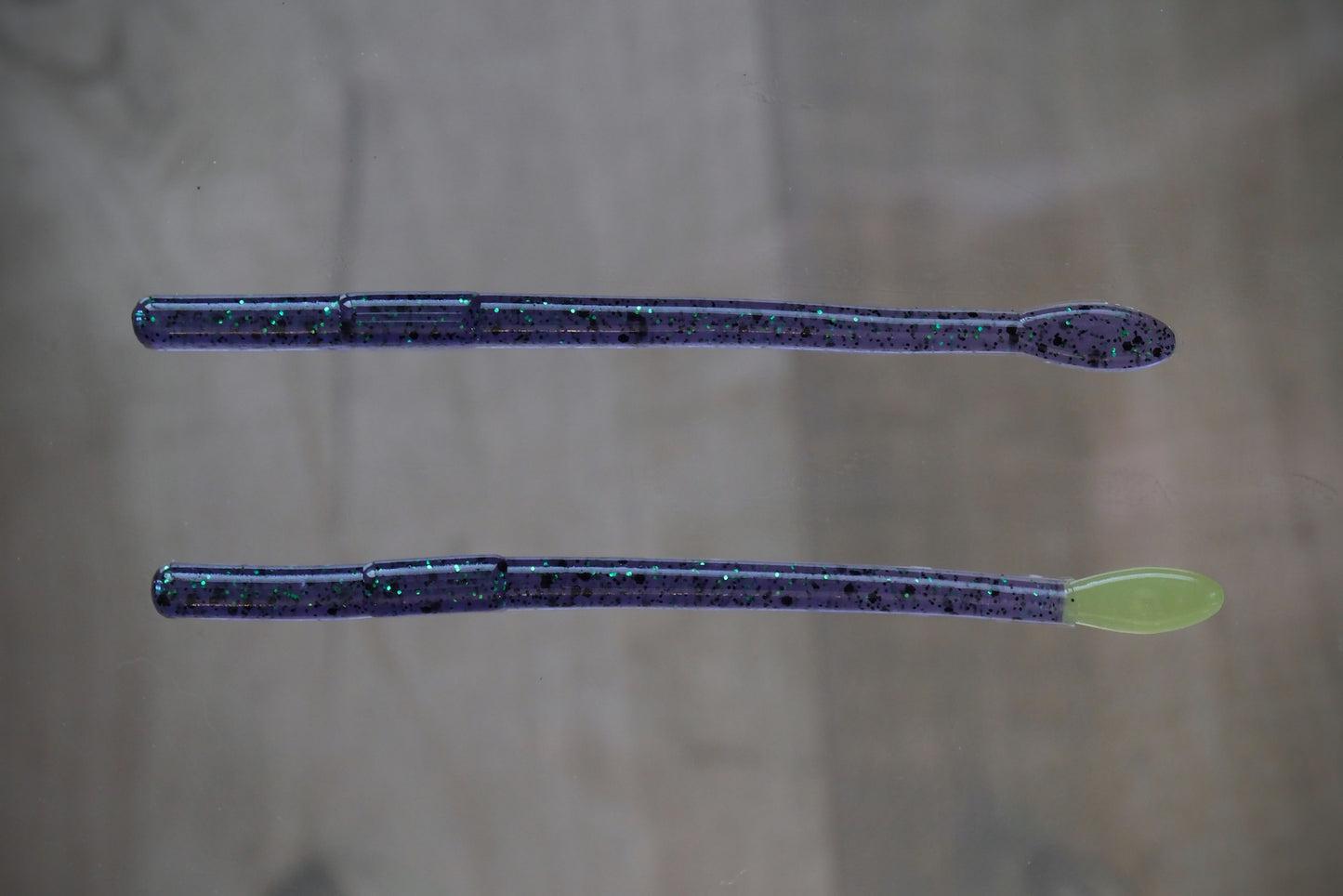 Two soft plastic fishing lures with a dark purple glitter body and a light green spoon tail, shown on a neutral gray background.