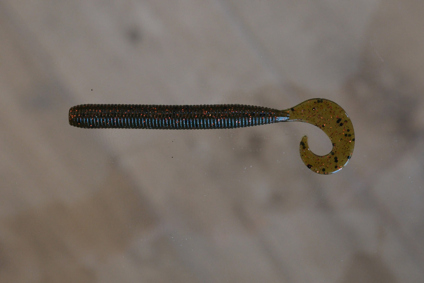 A soft plastic fishing lure shaped like a segmented worm with a curved hook tail, shown against a blurred beige background.