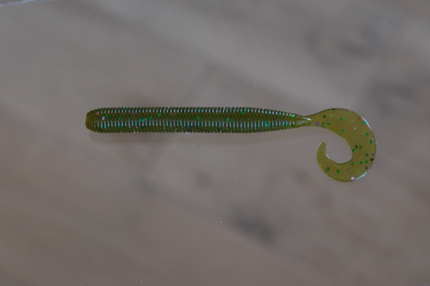 A green soft plastic worm with a curly tail, shown on a neutral beige surface.